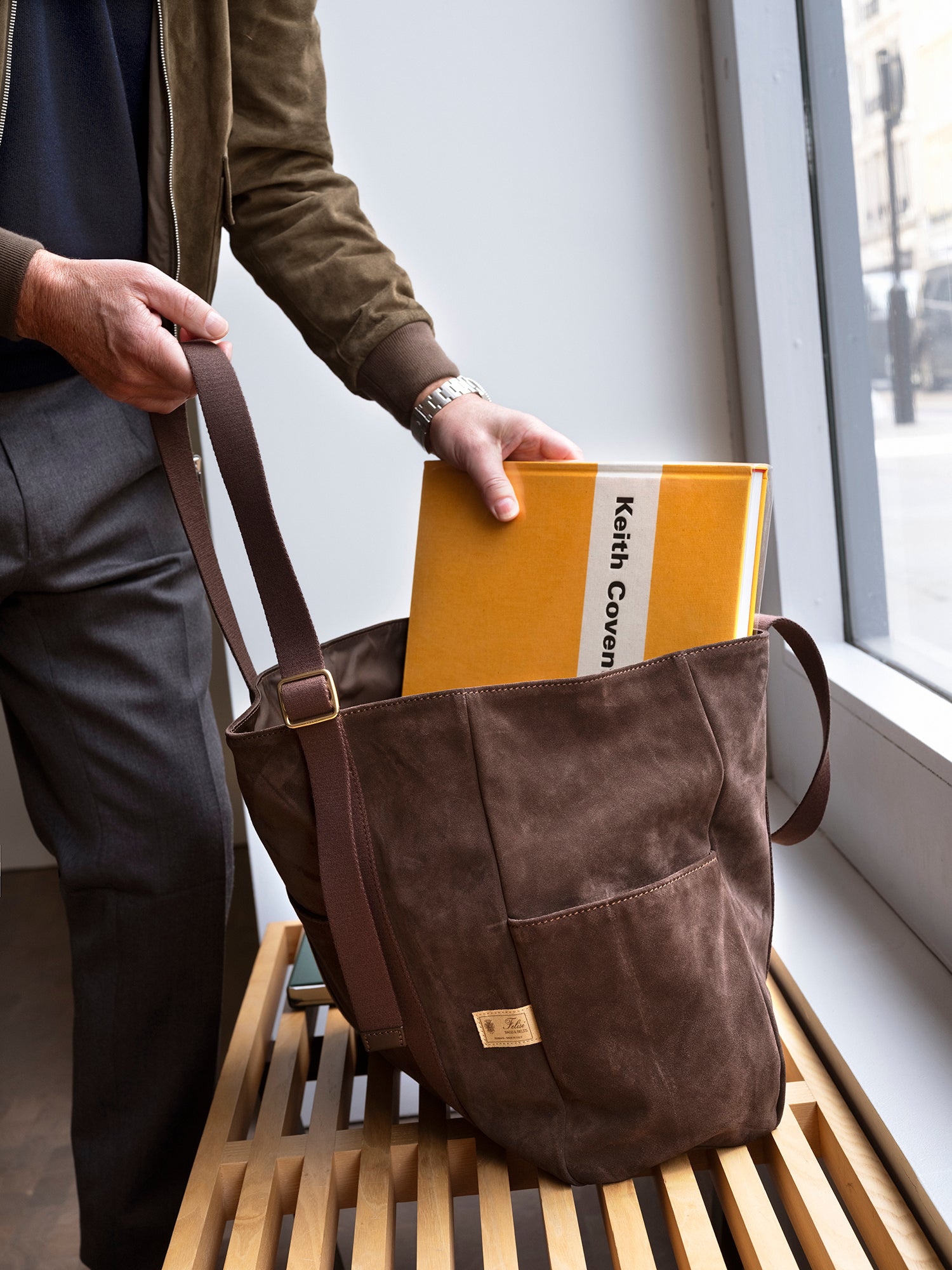 Person holding a brown leather bag with a yellow book titled 'Keith Cooper' by a window.