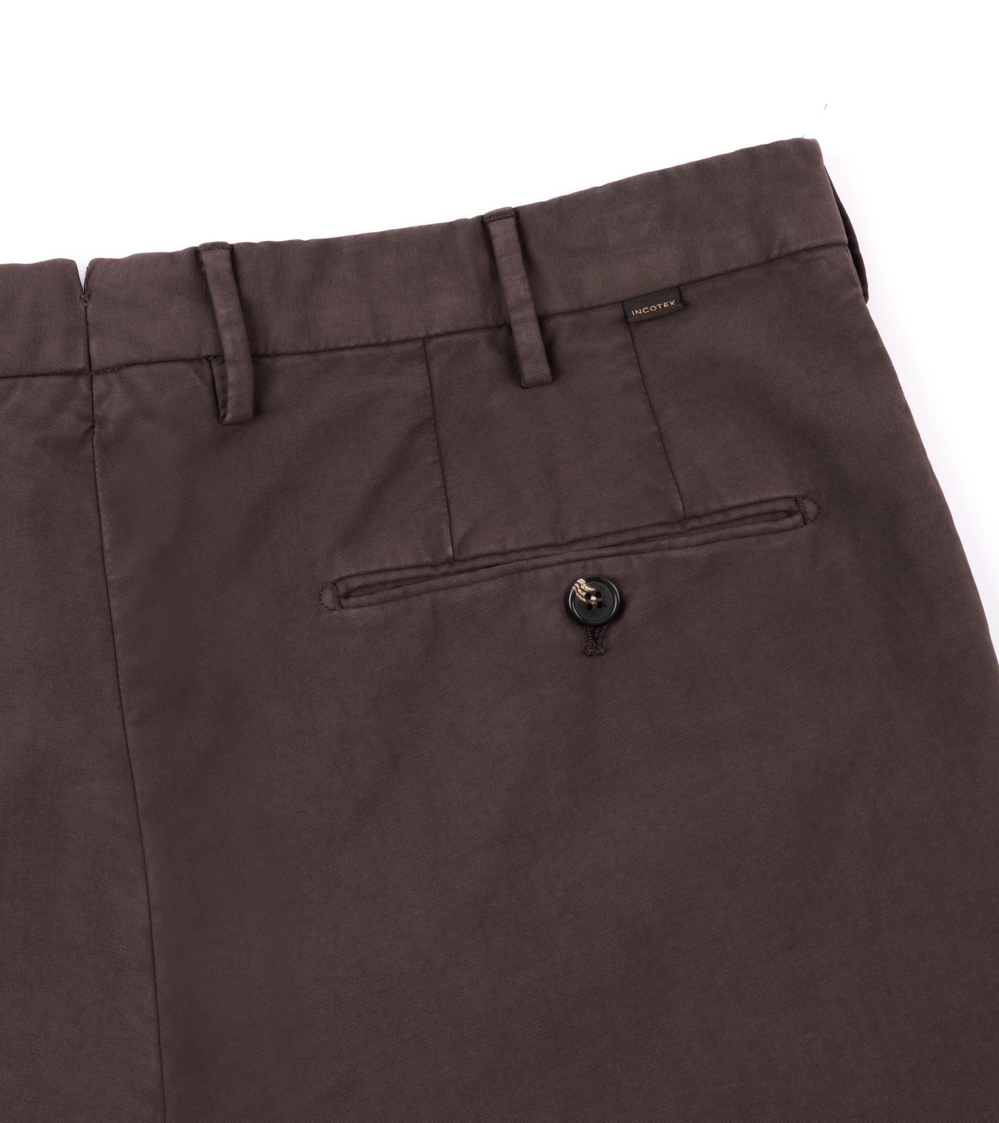 Close-up of brown trousers showing the waistband, belt loops, a rear welt pocket with a button, and a small woven brand tag near the back right side.
