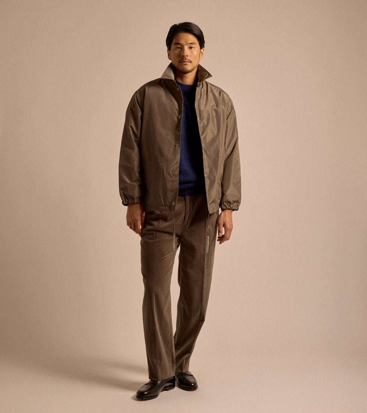 Model wearing Kaptain Sunshine Silk Poly Coach Jacket in Stone Grey. Front view, styled with Navy lambswool sweater, brown corduroy trousers and black leather loafers.