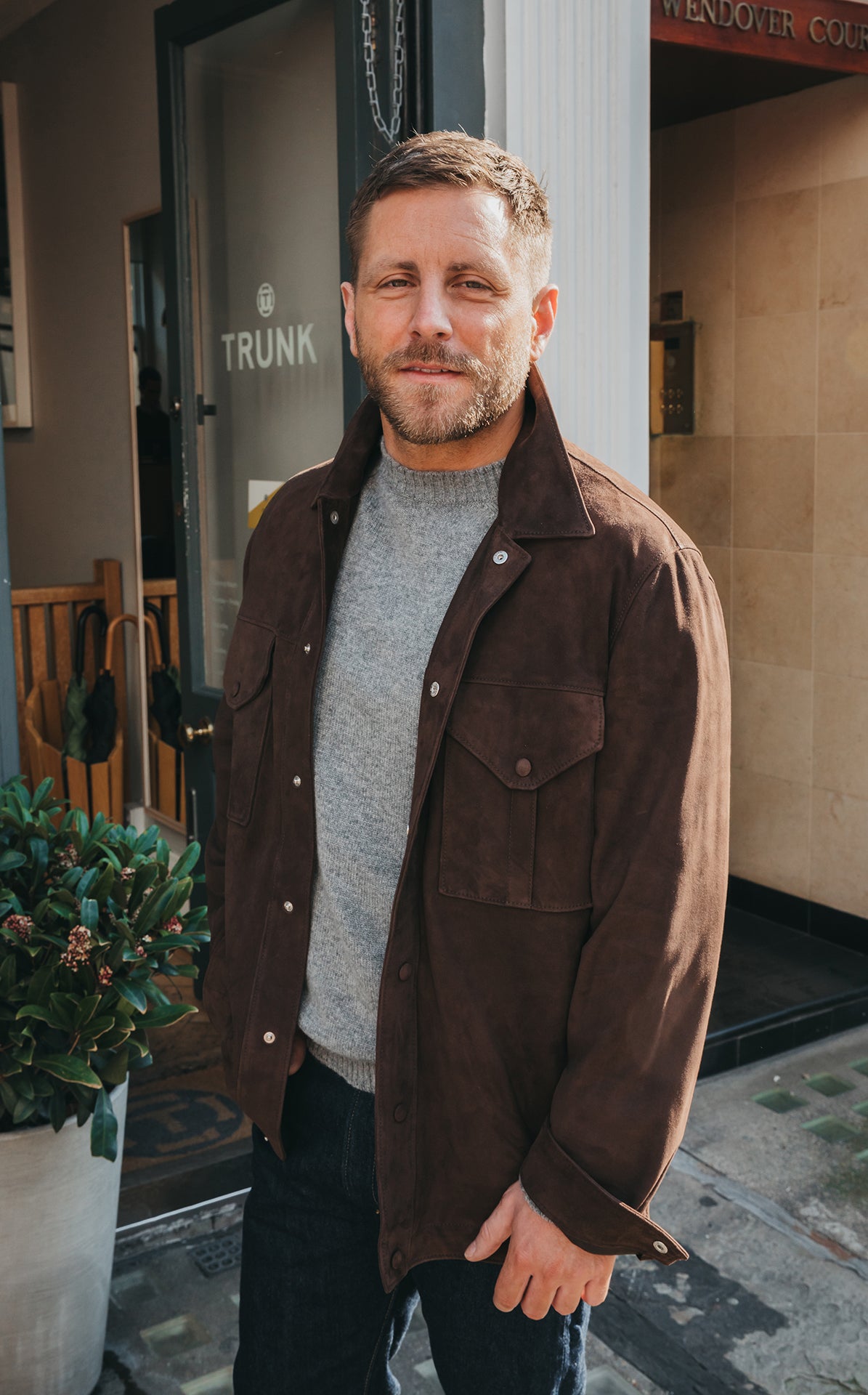 Man wearing a brown jacket and gray sweater standing outside a building with 'Trunk' visible.