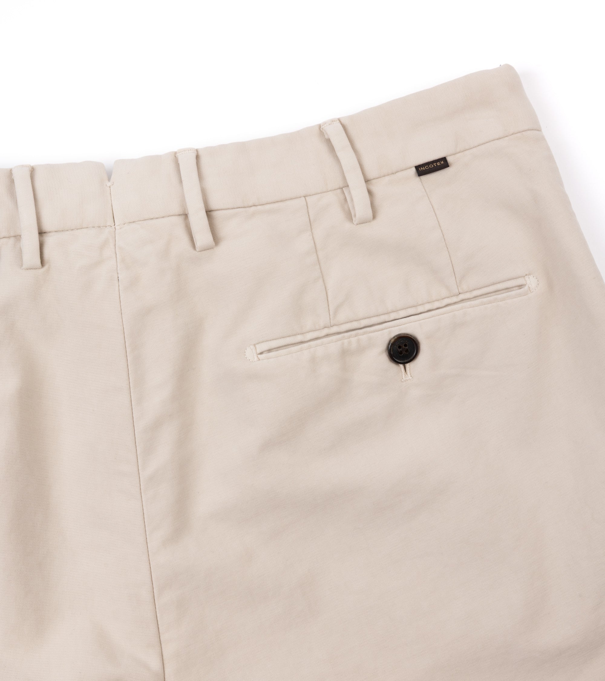Close-up of cream trousers showing the waistband, belt loops, a rear welt pocket with a button, and a small woven brand tag near the back right side.