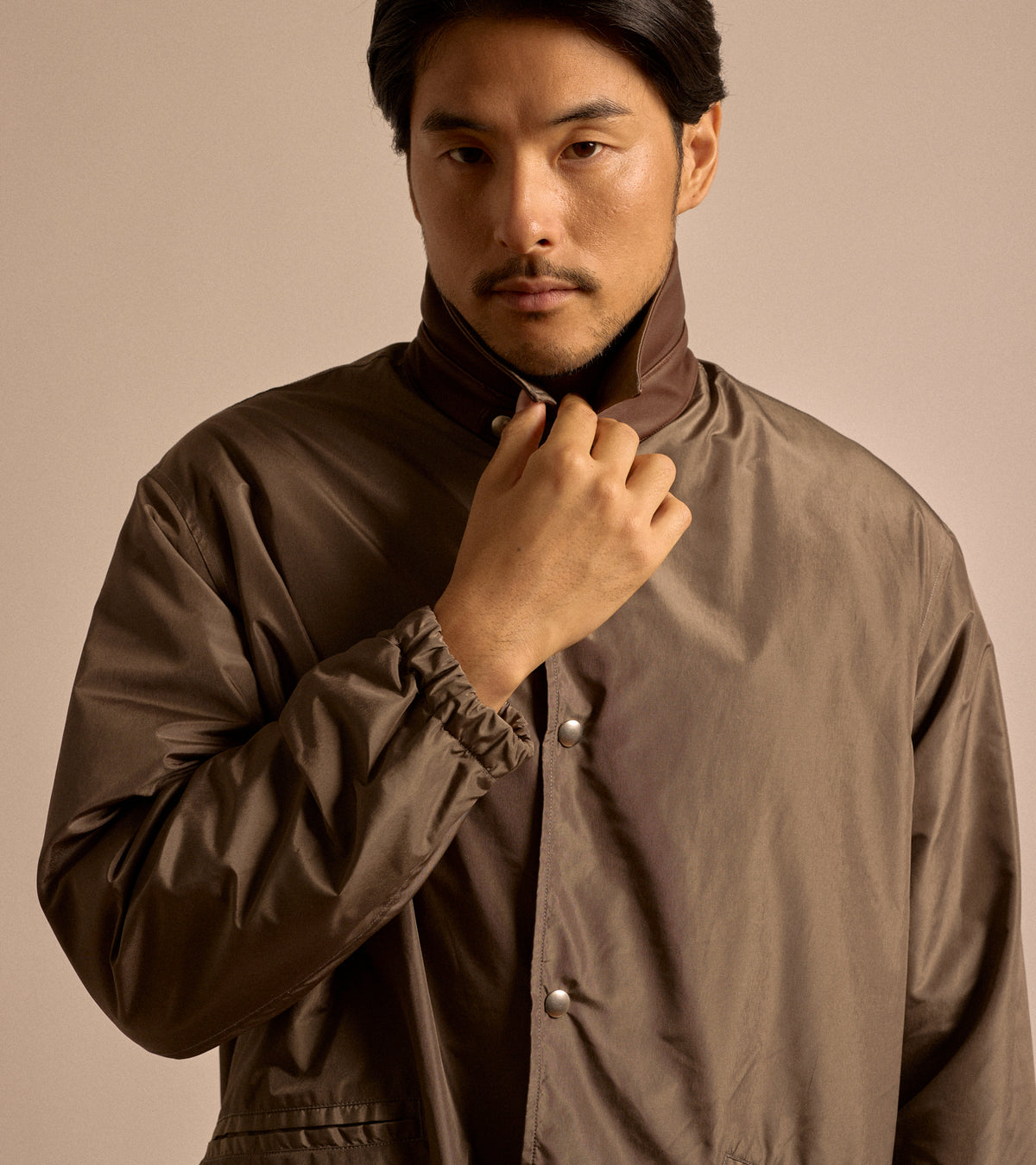 Model wearing Kaptain Sunshine Silk Poly Coach Jacket in Stone Grey.. Close-up shot of the model holding the leather collar up.