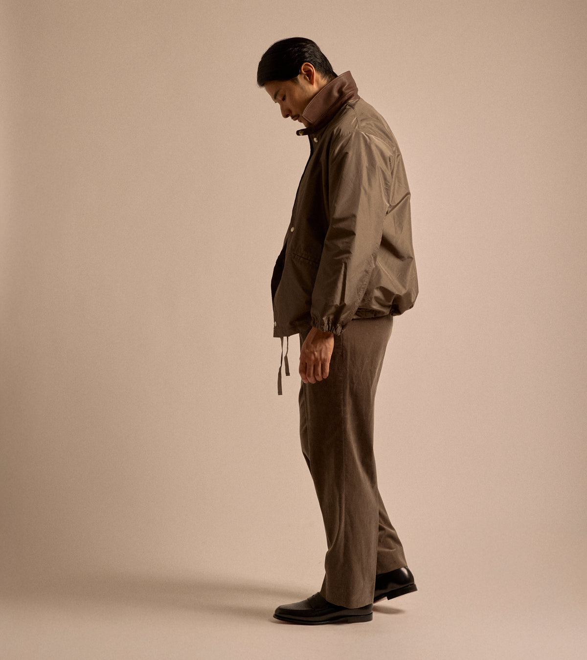 Model wearing the Kaptain Sunshine Silk Poly Coach Jacket in Stone Grey. Shown from a side view. Styled with brown corduroy trousers and black leather loafers.